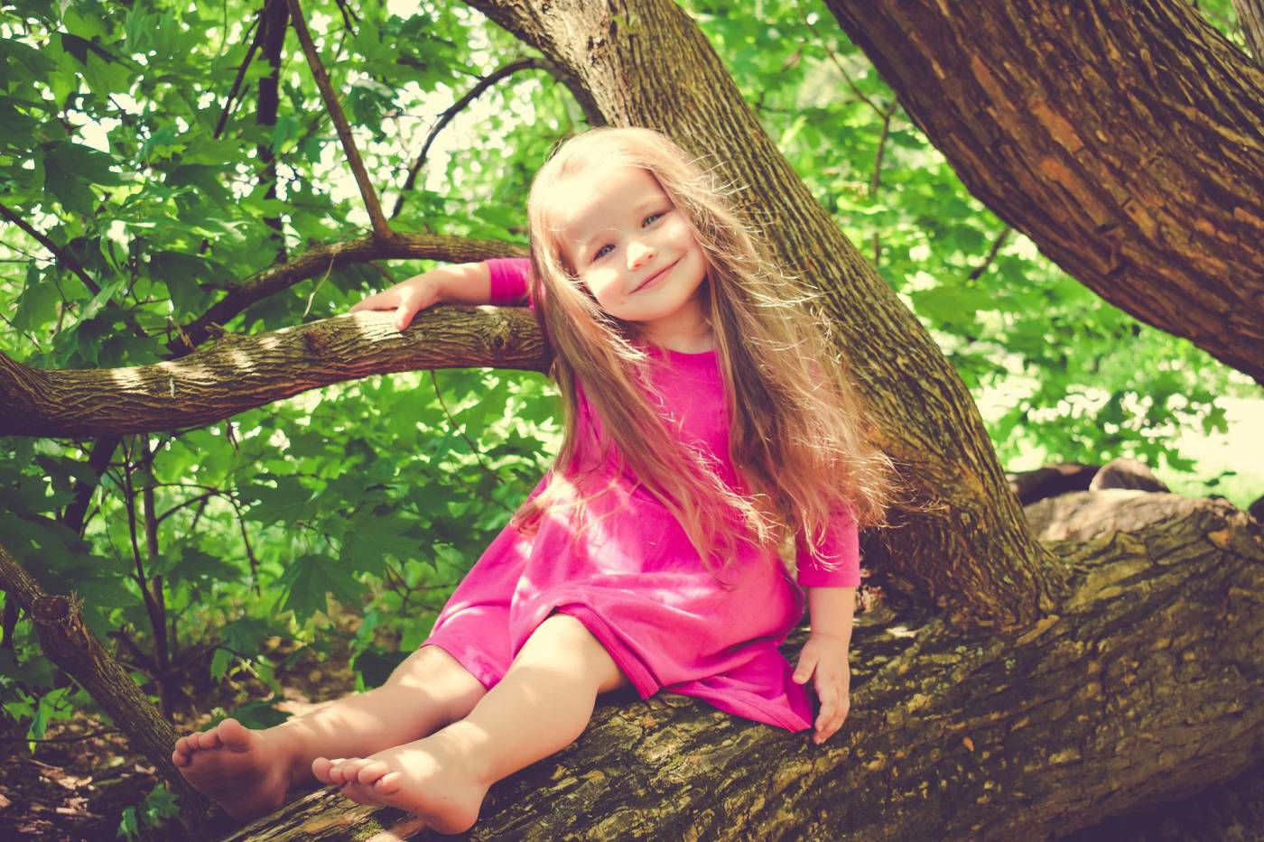 A joyful child with long hair sits on a tree branch, wearing a pink dress amidst vibrant green foliage.