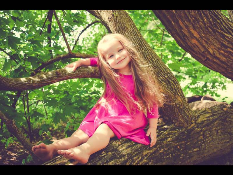 A joyful child with long hair sits on a tree branch, wearing a pink dress amidst vibrant green foliage.