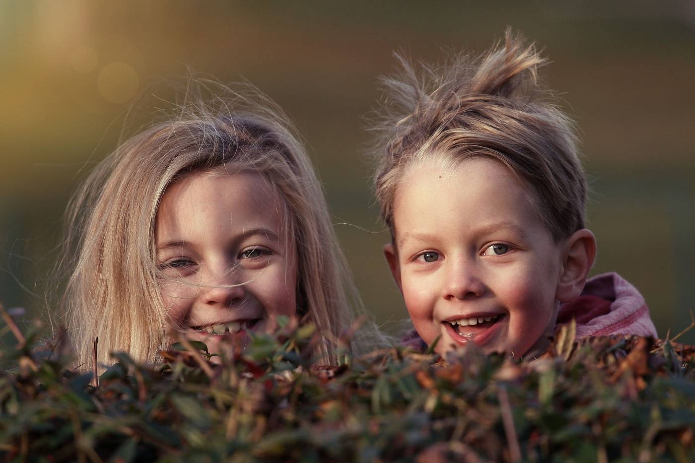 Two smiling children peek over a bush, with sunlight softly illuminating their joyful faces.