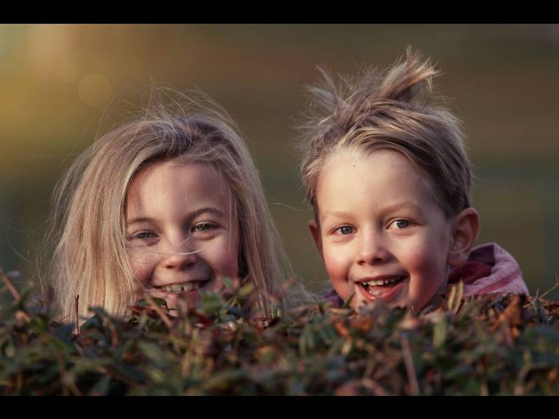 Two smiling children peek over a bush, with sunlight softly illuminating their joyful faces.