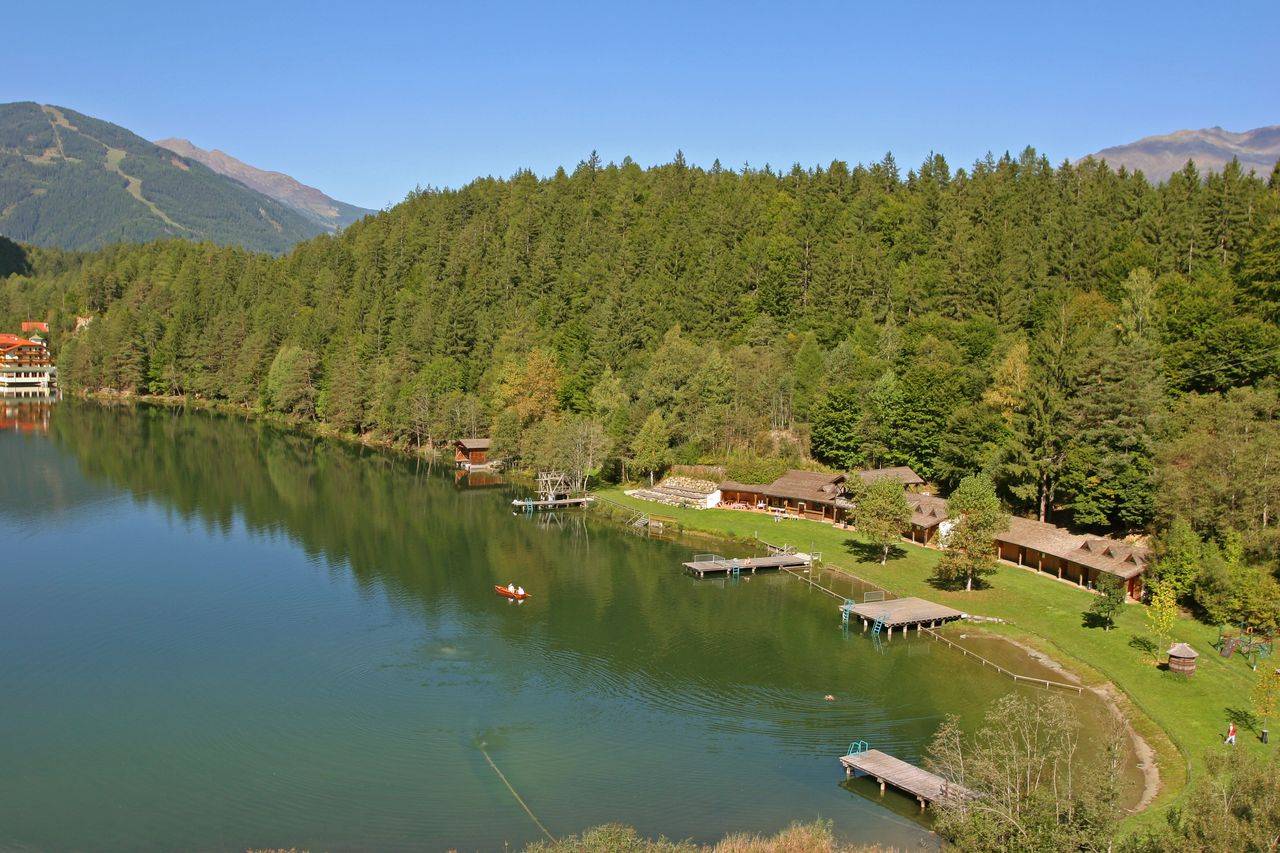 Ein ruhiger See umgeben von Wäldern, mit Bootsstegen und kleinen Häuschen im sonnigen Gelände.