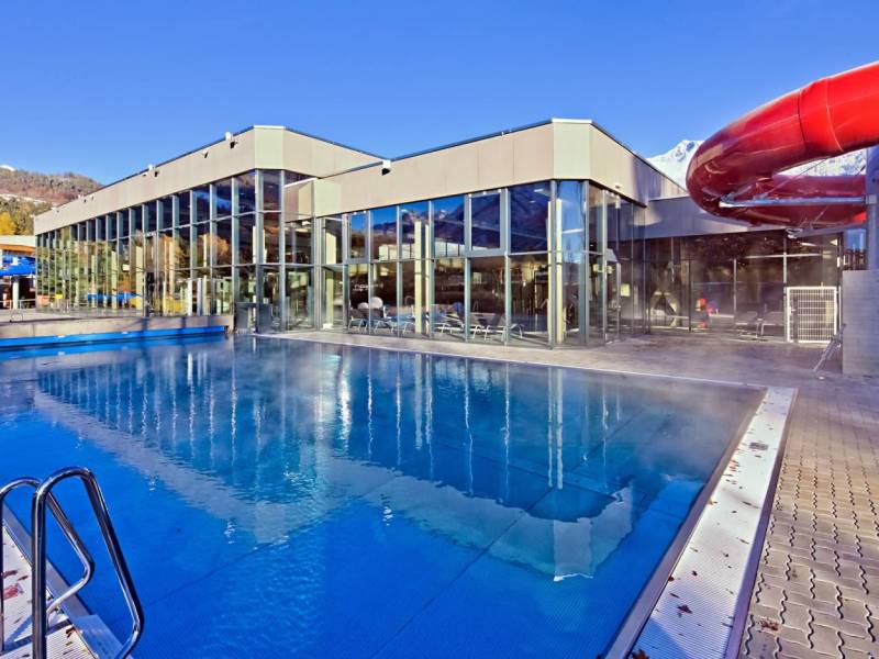 Ein modernes Schwimmbad mit Glasfront und einer roten Wasserrutsche bei klarem Himmel und dampfendem Wasser.