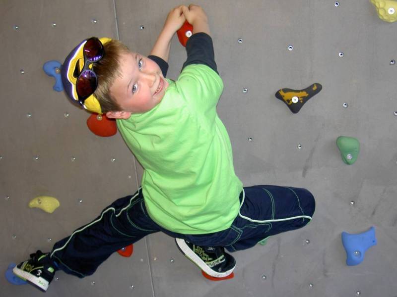 Ein Junge klettert fröhlich an einer Kletterwand, trägt ein grünes Shirt, Sonnenbrille und hat Spaß.
