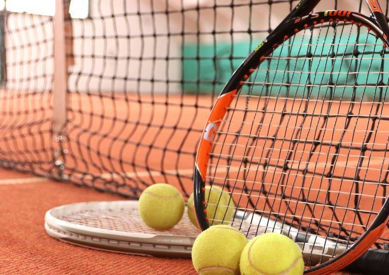 Tennisplatz mit zwei Schlägern und mehreren gelben Tennisbällen vor einem Netz in einer Halle.