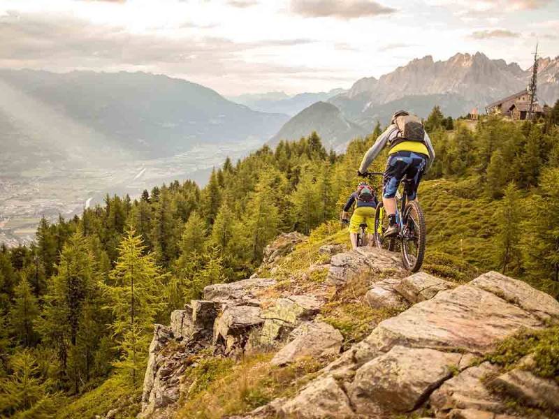  mountainbikende Fahrer auf einem felsigen Pfad mit Blick auf ein Tal und Berge im Hintergrund.