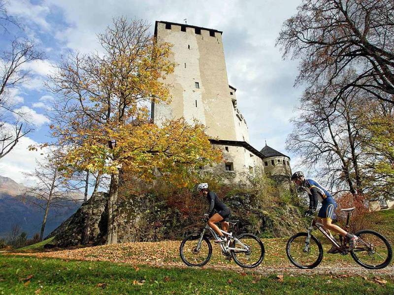 Zwei Mountainbiker fahren an einer alten Burg vorbei, umgeben von bunten Herbstbäumen und malerischer Landschaft.
