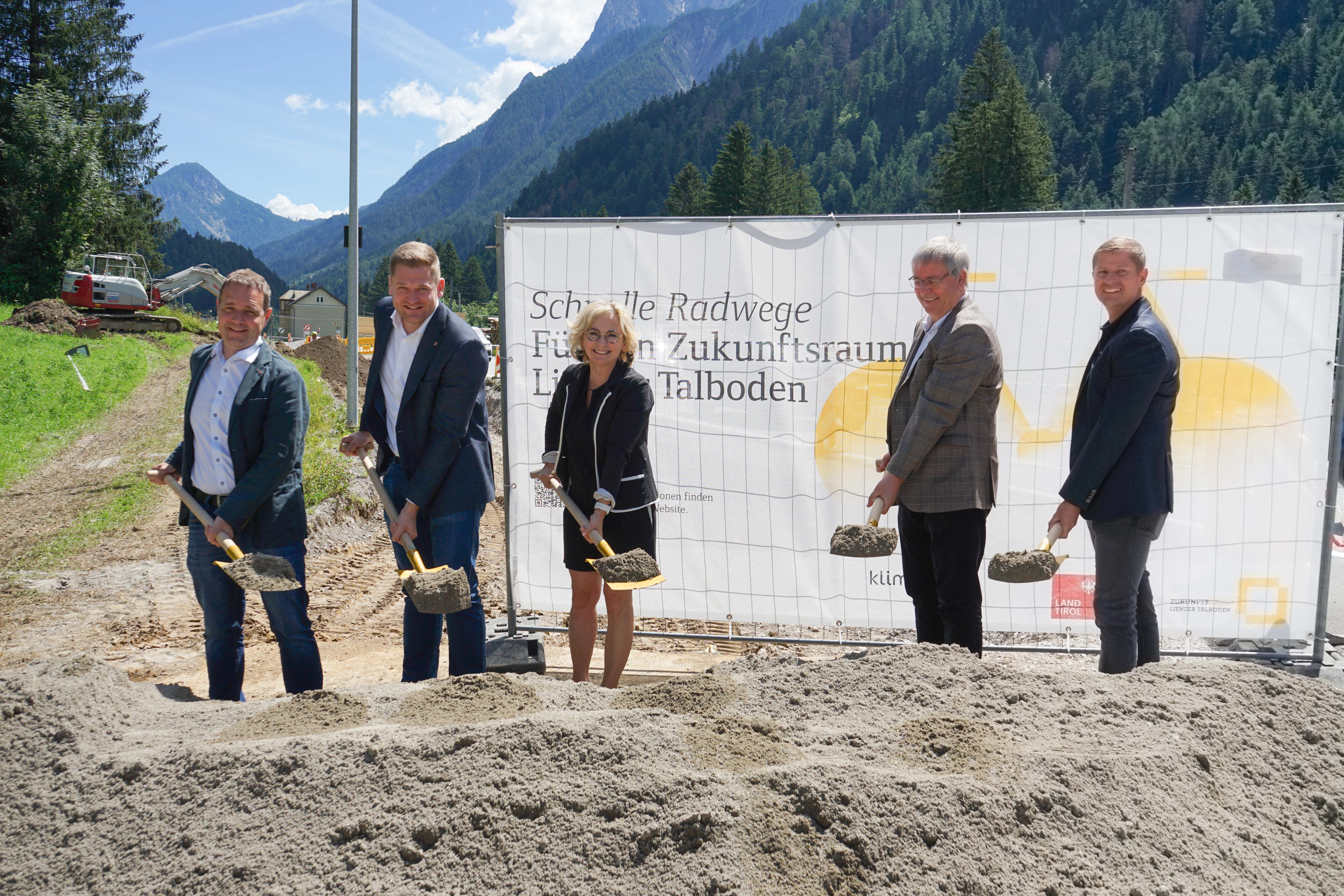 Spatenstich für neue Radwege in einer alpinen Umgebung, Teilnehmer stehen lächelnd hinter einem Sandhaufen.