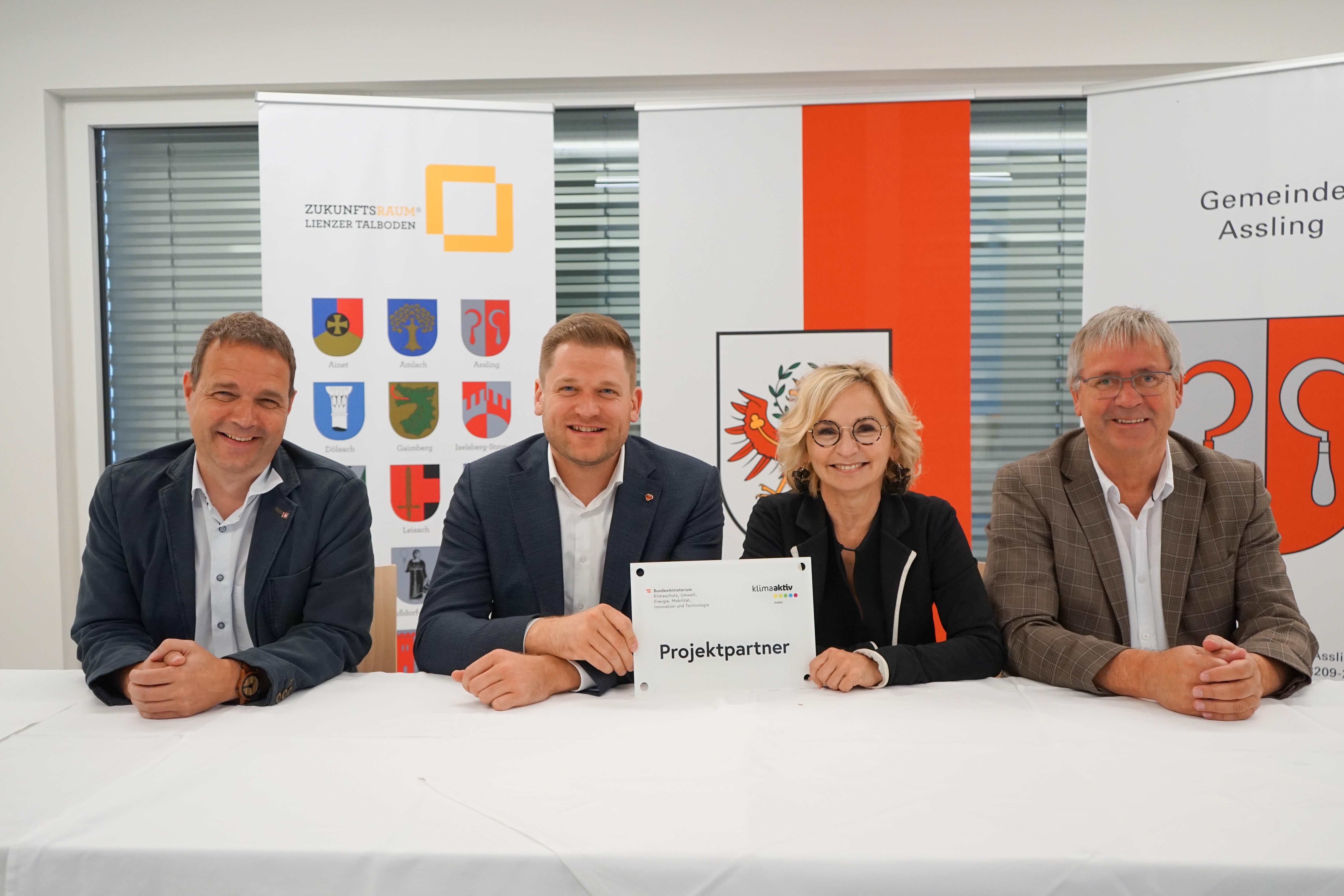 Vier Personen sitzen an einem Tisch und halten ein Schild mit der Aufschrift „Projektpartner“ in einer Besprechung.