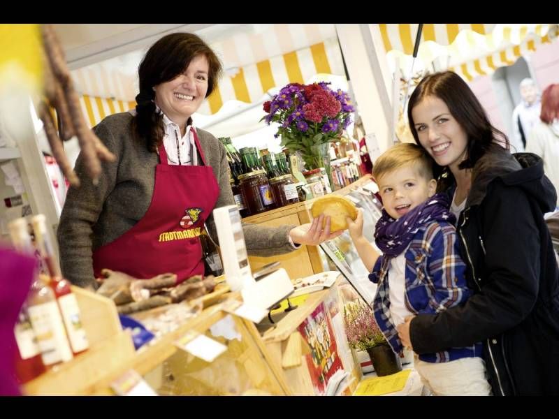 Eine junge Mutter kauft mit ihrem Sohn am Stadtmarkt einen Krapfen von einer sympathisch lächelnden Verkäuferin.