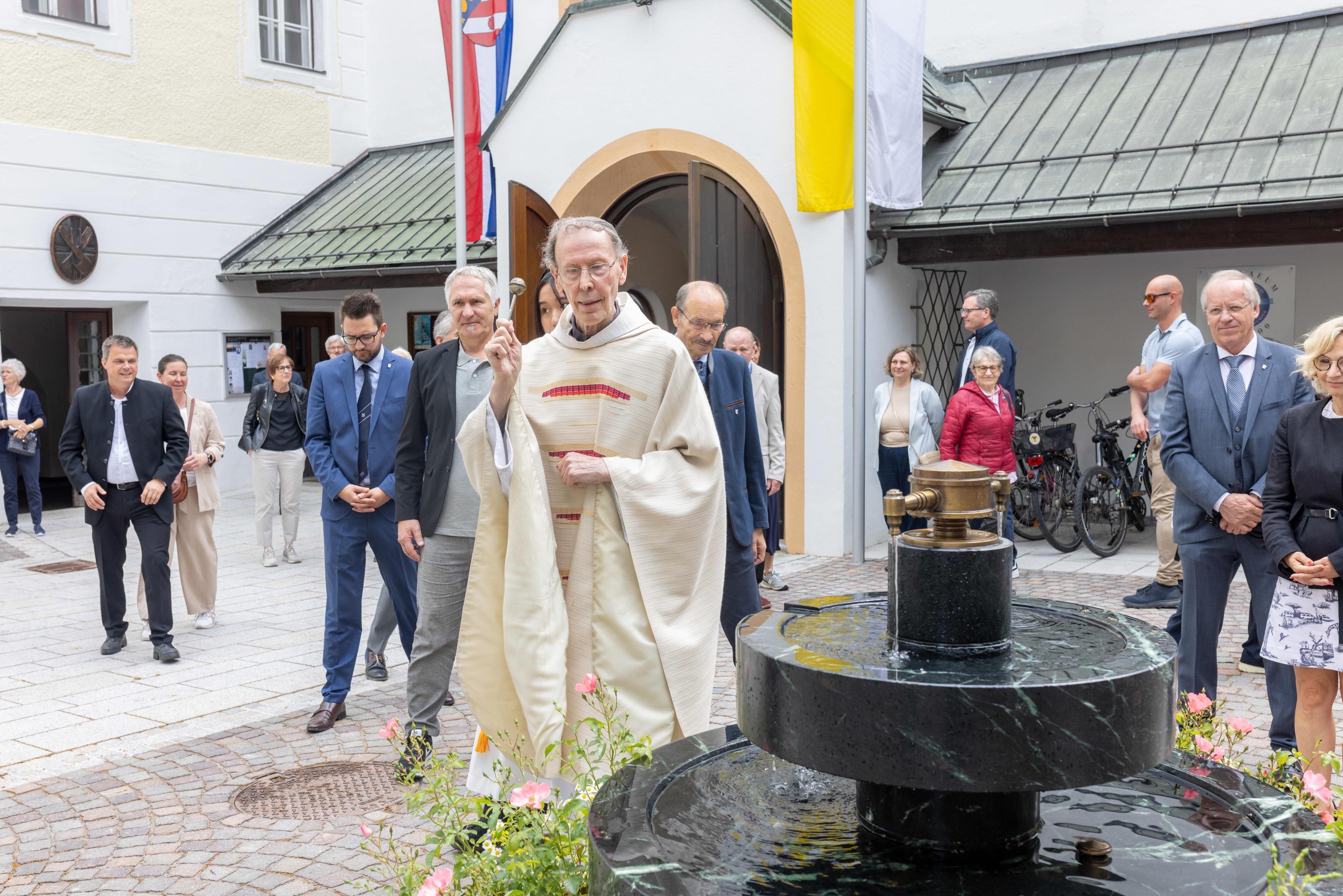 Eine Person in religiöser Kleidung führt eine Zeremonie mit Wasserfontäne vor Zuschauern durch.