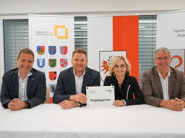 Vier Personen sitzen an einem Tisch, halten ein Schild mit "Projektpartner" und präsentieren ihre Zusammenarbeit.