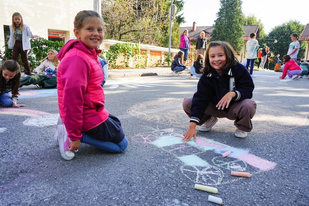 Zwei Mädchen zeichnen mit Kreide auf dem Asphalt, umgeben von anderen Kindern. Fröhliche Stimmung im Freien.