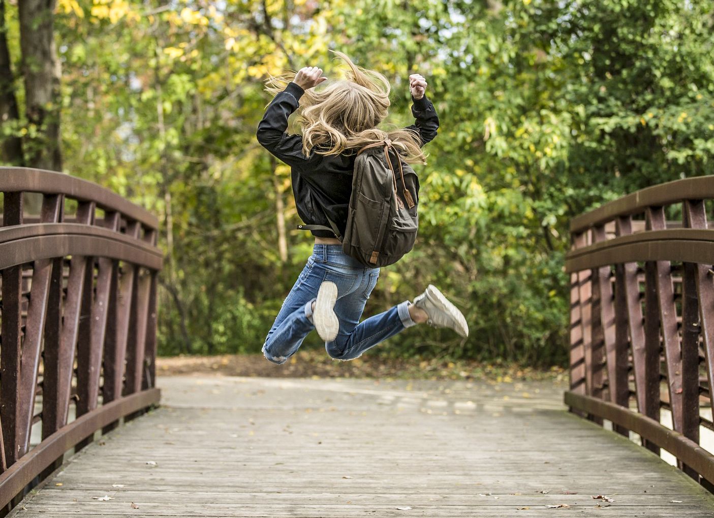 Ein Mädchen springt fröhlich auf einer Brücke in einem herbstlichen Park mit grünem Hintergrund.