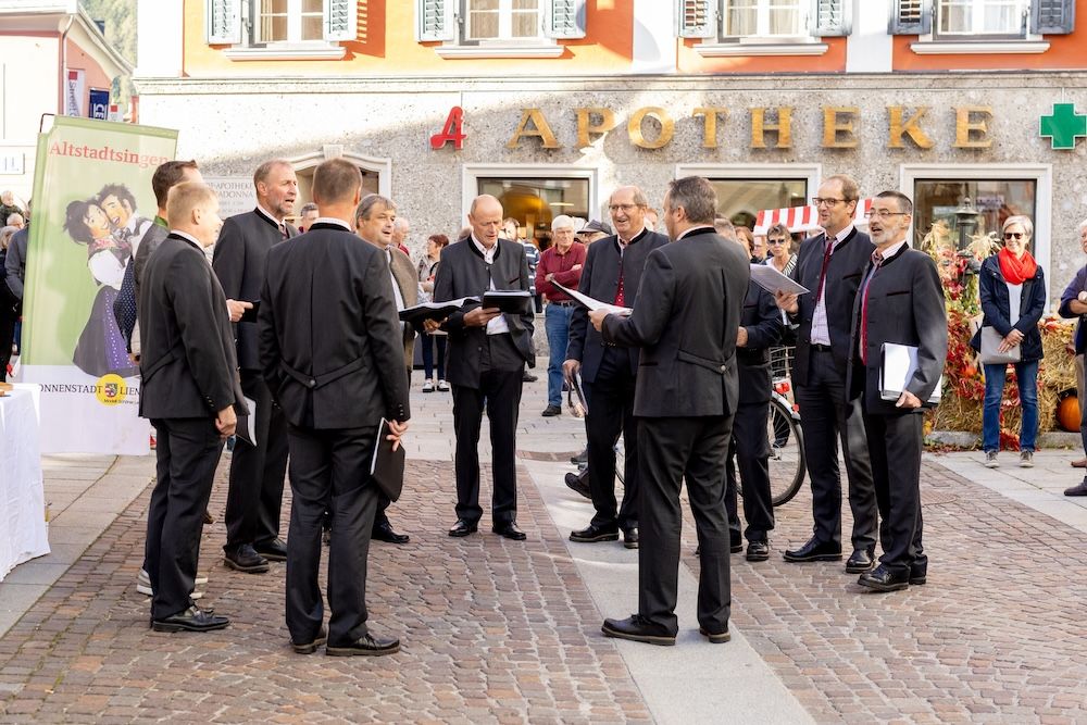 Eine Gruppe von Sängern singt in einer Stadt, umgeben von Zuschauern und bunten Dekorationen.