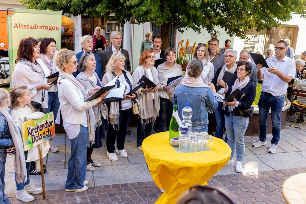 Ein Chor singt im Freien, umgeben von Zuschauern. Es ist eine fröhliche, gesellige Atmosphäre.